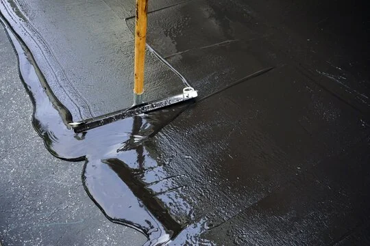 Worker applying asphalt sealcoating on a paved surface using a spreader tool.